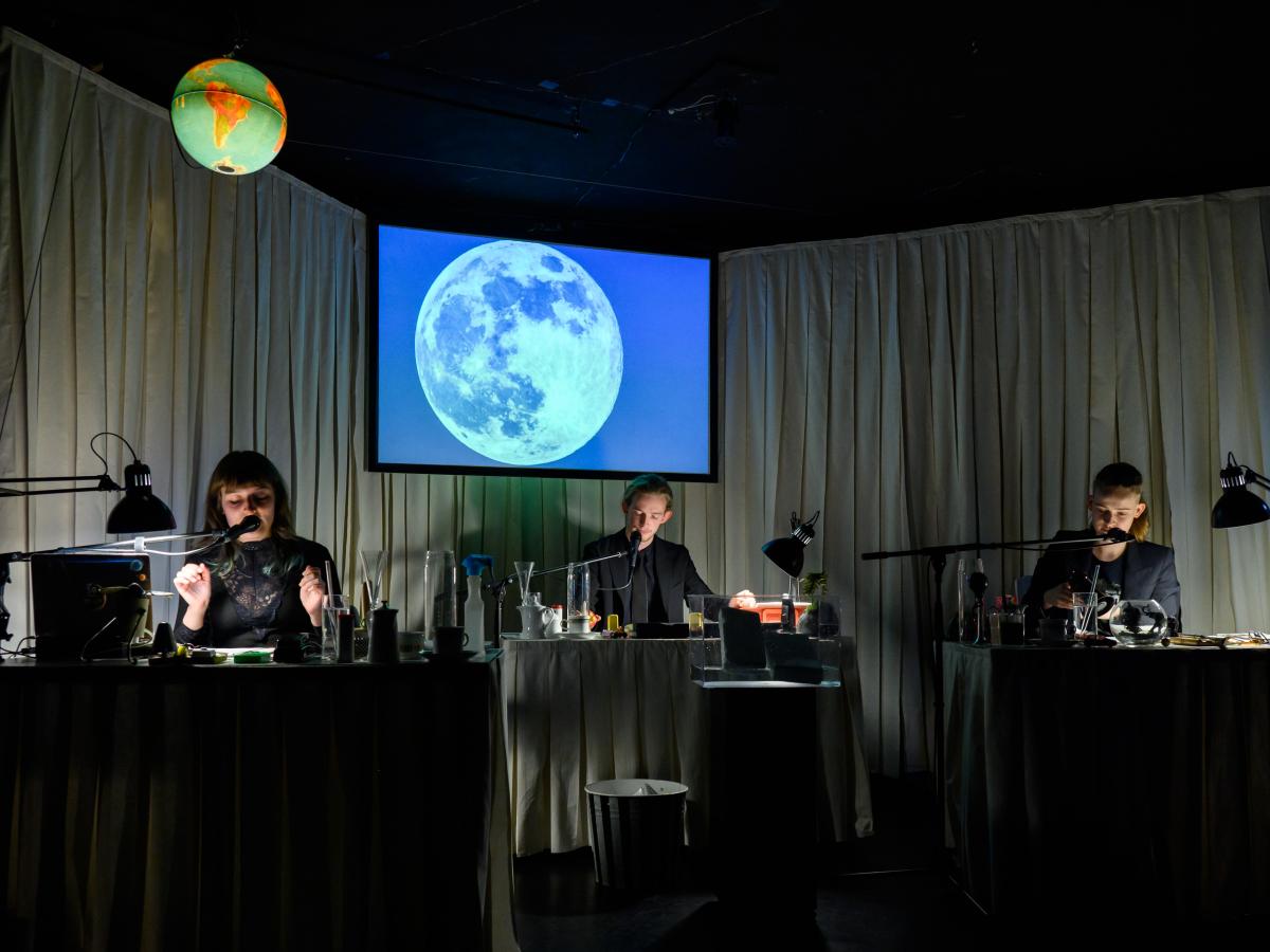 drei menschen an Tischen mit Mikrophonen. Im Hintergrund ein Planet auf großem Bildschirm.