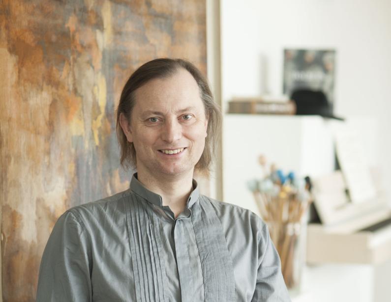 Klaus Dreher vor einem abstrakten, in Bastellfarben gehaltenen Bild auf einem grauen Sessel sitzend, im Hintergrund ein weißlackiertes Klavier.ier im Hintergrund