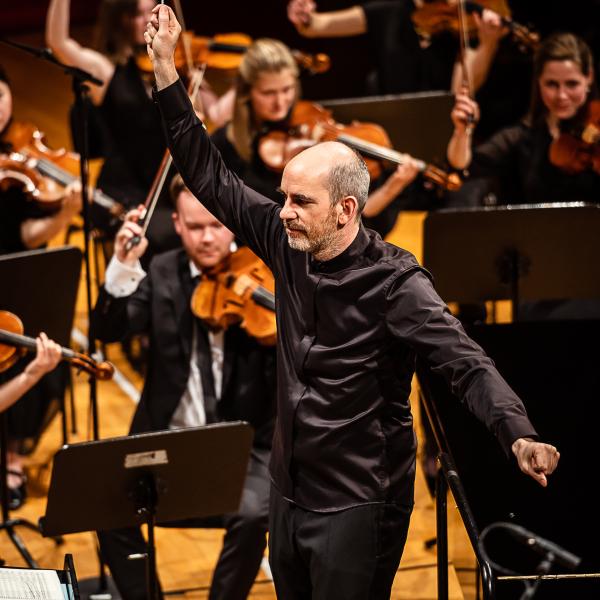 Der Dirigent Rasmus Baumann mit einer ausladenden Geste. Mit freudigen Gesichtsausdruck inmitten des Orchesters.