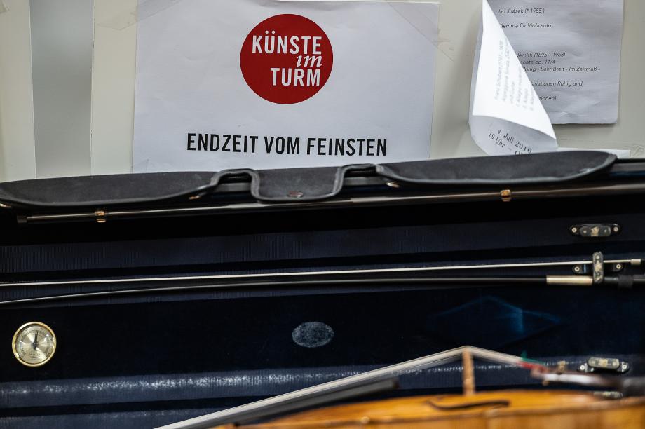 Ein aufgeschlagener Geigenkasten, der vor einem Konzertplakat mit dem Titel "Endzeit vom Feinsten" positioniert ist.