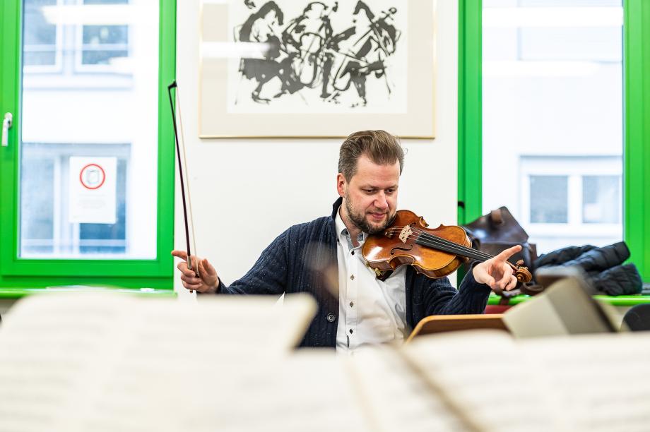 Violinprofessor Martin Funda in der Frontalansicht. Die rechte Hand mit dem Geigenbogen ist weit ausgestreckt.