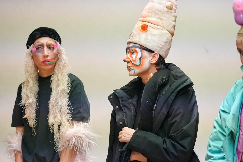 zwei Frauen: eine, blonnde Wollhaare, pinke Federwimpern und Regenbogenmund, die andere hat ein orange-schwarz-weiß geschminktes Gesicht und eine hohe Kopfbedeckung mit Muscheln auf.