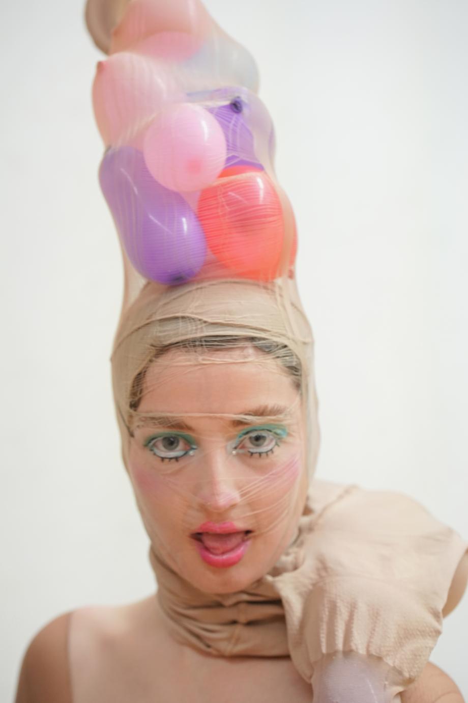 Frau in Nahaufnahme, offenem Mund, die Augen comic-mäßig grün geschmikt, mit aufgezeichneten Wimpern. Auf dem Kopf trägt sie bunten Ballons, die von einer Feinstrumpfhose, welche ihr auch über das Gesicht geht, gehalten werden.