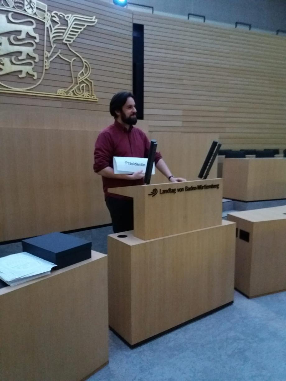 Mann im Landtag Baden-Württemberg hinter Rednerpult und hät ein Schild in der Hand, auf dem Präsidentin steht.